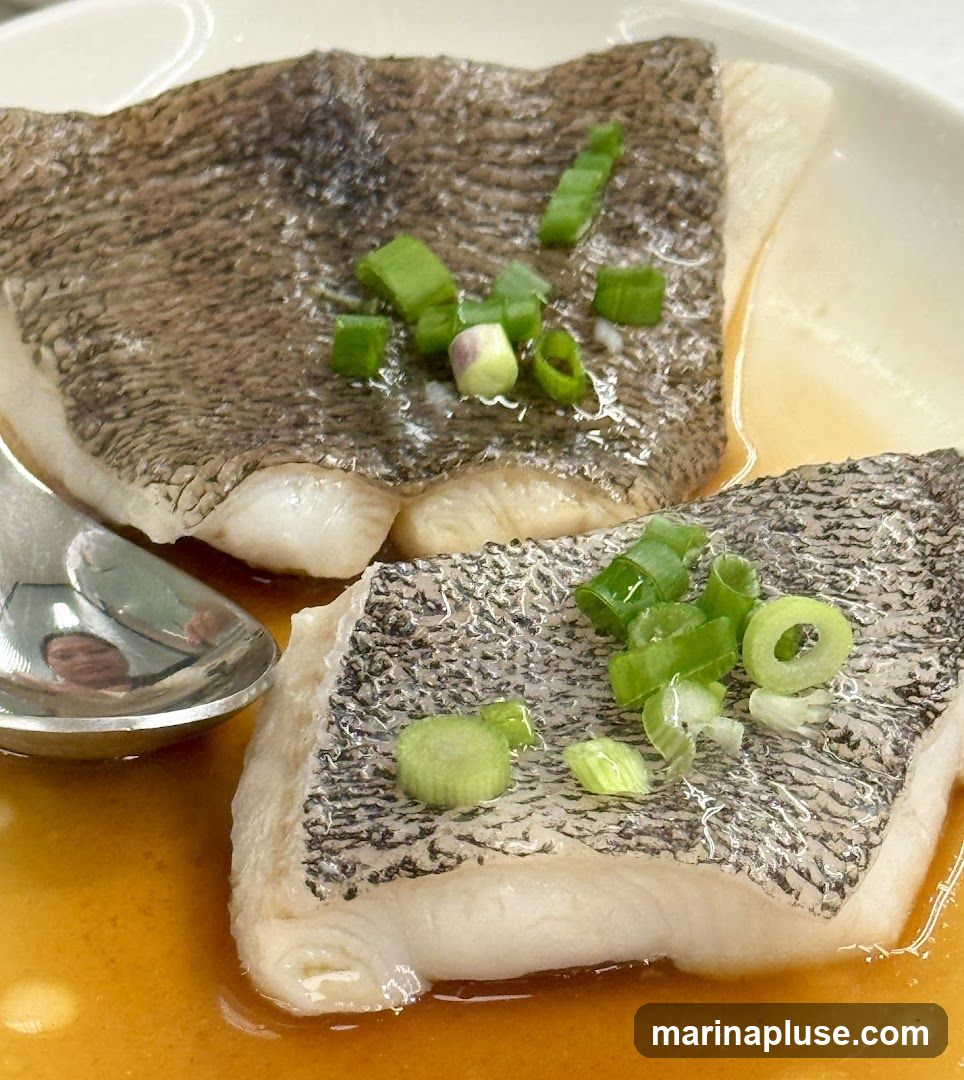Two pieces of steamed fish fillets with crispy skin, garnished with chopped green onions, served in a light brown sauce. A spoon is visible on the left side of the dish, reflecting a person's face.