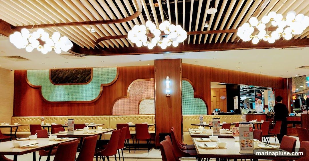 The image shows the interior of a restaurant with tables and chairs set up for dining. The decor features wooden paneling, decorative wall accents, and modern chandeliers. Several tables are visible, each set with bowls and chopsticks. In the background, a staff member is standing near a counter, and promotional materials are displayed on some tables.