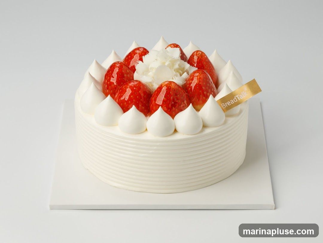 A round white cake with whipped cream dollops around the edge, topped with fresh strawberries and a small cluster of white flowers in the center. A gold ribbon with "BreadTalk" branding is placed on top right. The cake sits on a square white plate against a plain white background.
