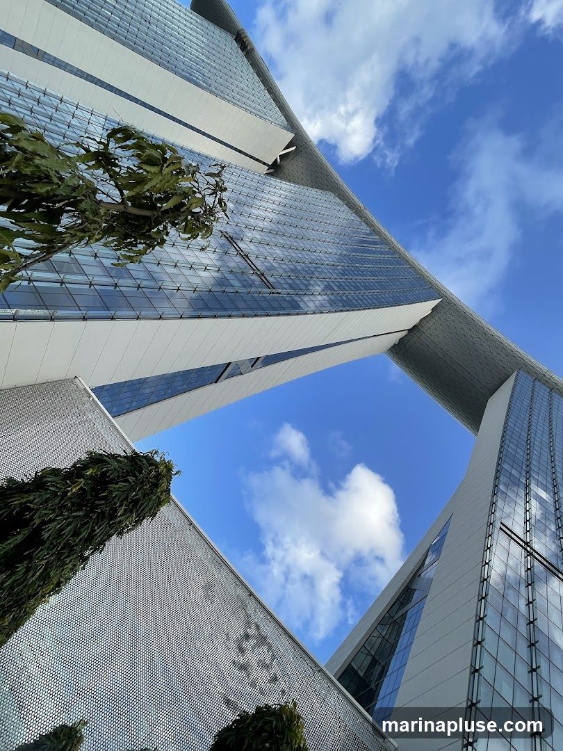 A low-angle shot of a modern architectural complex featuring sleek, glass-covered buildings against a bright blue sky with scattered clouds. The buildings have a distinctive, angular design, with one section appearing to bridge over an open space. Lush green trees are visible in the foreground, adding a natural element to the urban landscape. The reflective surfaces of the buildings mirror the sky and surrounding structures.