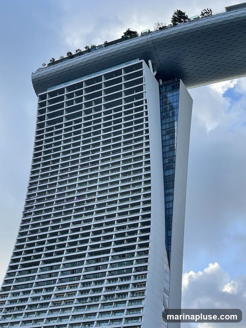 A close-up, low-angle shot of the Marina Bay Sands hotel in Singapore. The iconic building features a distinctive curved facade with numerous balconies and a large, boat-shaped structure on top, which is adorned with trees and greenery. The sky above is partly cloudy.