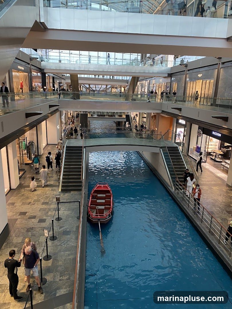 An interior view of a multi-level shopping mall featuring a canal with a red gondola. People are seen walking on the walkways and bridges above and around the canal. Shops with glass storefronts are visible on multiple levels.