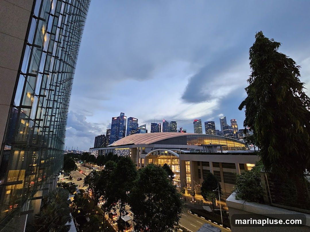 A view of a modern cityscape at dusk, with tall buildings and a large domed structure illuminated. A glass-fronted building is on the left, reflecting the sky and lights. Trees and a street with traffic are visible in the foreground.