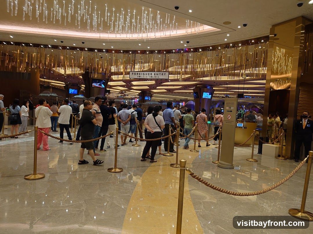 A crowd of people are queuing inside a large, opulent building, likely a casino. The ceiling features elaborate chandeliers, and the walls are decorated with golden accents. A sign overhead reads "FOREIGNER ENTRY" with accompanying text in what appears to be Chinese characters. Ropes and stanchions guide the queue. A security guard in uniform stands to the right.