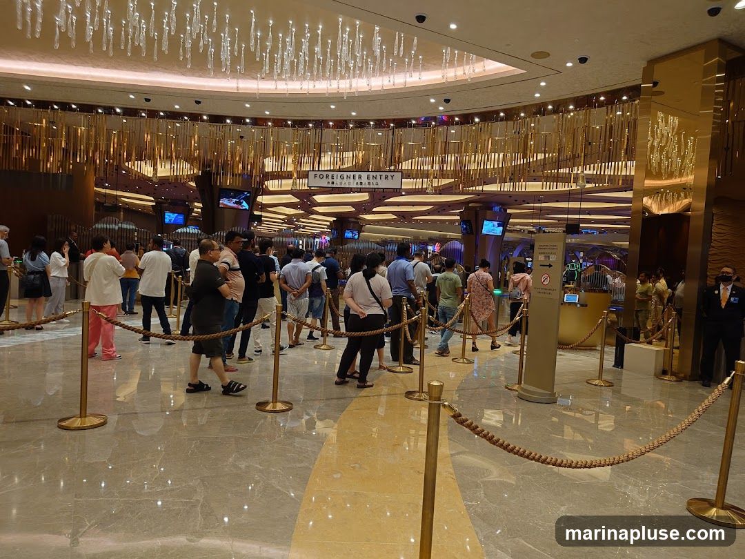 A crowd of people are queuing inside a large, opulent building, likely a casino. The ceiling features elaborate chandeliers, and the walls are decorated with golden accents. A sign overhead reads "FOREIGNER ENTRY" with accompanying text in what appears to be Chinese characters. Ropes and stanchions guide the queue. A security guard in uniform stands to the right.
