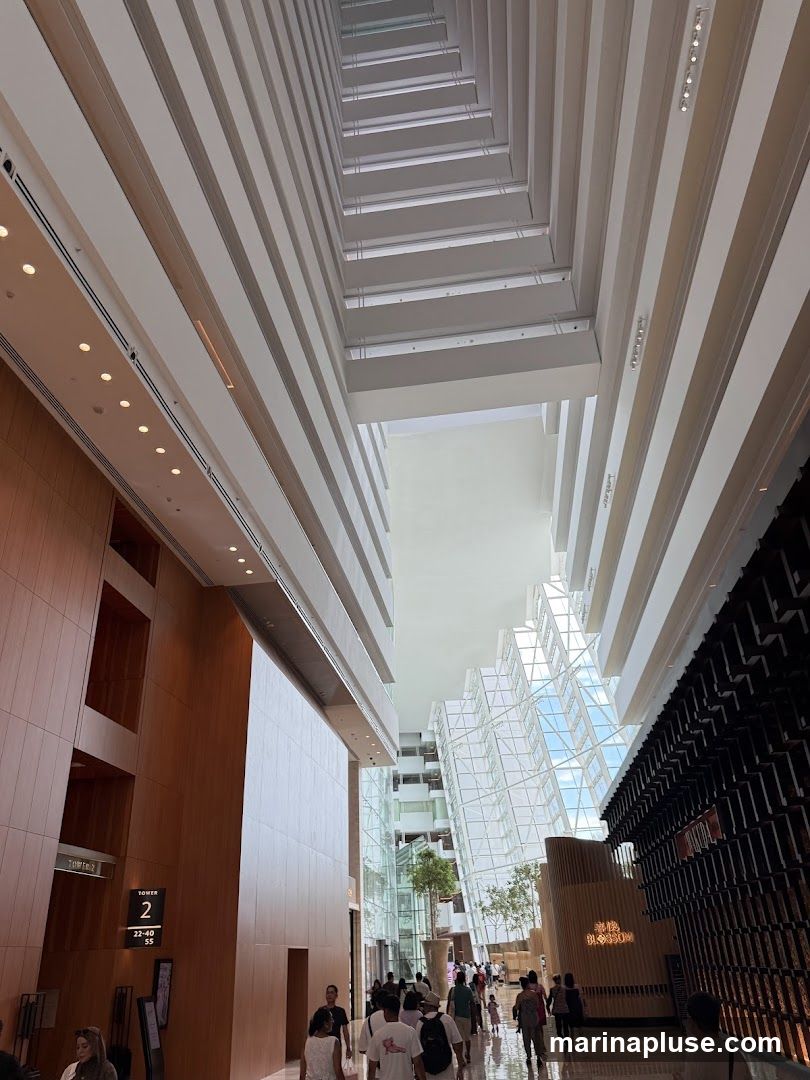 A wide-angle shot looking up into a modern atrium with multiple levels and a glass-paneled ceiling. The architecture features clean lines and a mix of wood paneling and dark, textured walls. People are walking through the spacious interior, and there are some trees and decorative elements visible. A sign indicates "TOWER 2" with floor numbers.