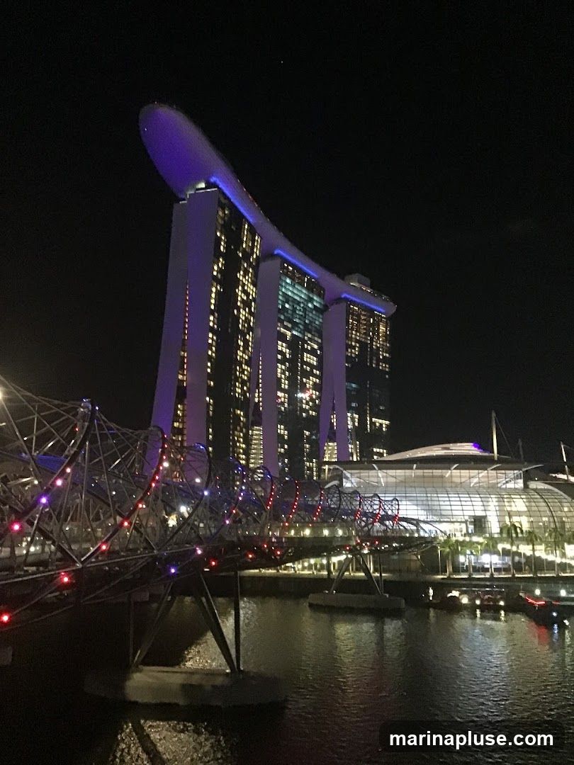A nighttime view of the Marina Bay Sands hotel and the Helix Bridge in Singapore. The hotel is illuminated with purple and white lights, and its iconic boat-shaped rooftop is visible. The Helix Bridge, a pedestrian bridge, is lit with colorful lights. The Singapore River flows in the foreground, reflecting the lights of the city.