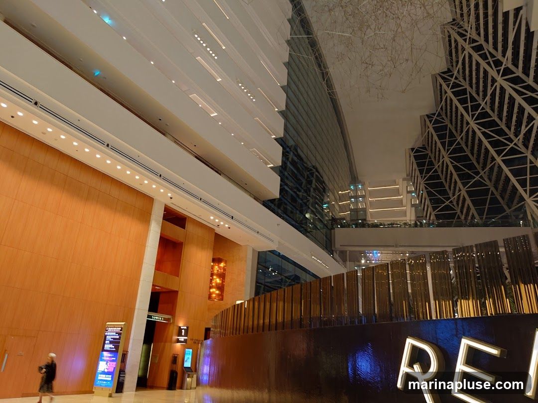A wide-angle shot of a modern interior space with a high ceiling and multiple levels. The walls are clad in warm-toned wood panels, contrasting with the sleek white architectural elements and glass facades. A prominent feature is a dark, reflective surface in the foreground with illuminated letters spelling out "REI". The background shows a complex geometric structure and a sky-like ceiling with intricate wire art. Digital signage and directional signs are visible on the left side.
