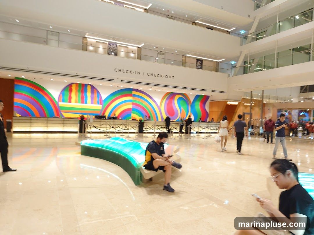 A wide shot of a hotel lobby featuring a long reception desk with a vibrant, abstract art installation above it. The art consists of colorful curved and vertical stripes. Text above the desk reads "CHECK-IN / CHECK-OUT". Several people are visible in the lobby, some interacting with the reception desk, others seated or walking through the space. A modern, illuminated bench is positioned in the foreground. The lobby has a polished marble floor and a multi-level architectural design with balconies overlooking the main area.