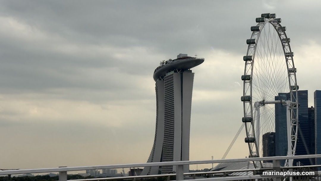 A wide shot of the Singapore skyline under a cloudy sky. Prominently featured are the iconic Marina Bay Sands hotel, with its distinctive boat-shaped top, and the Singapore Flyer observation wheel. Other modern skyscrapers are visible in the background. A white railing runs across the foreground, suggesting the photo was taken from a bridge or elevated walkway.
