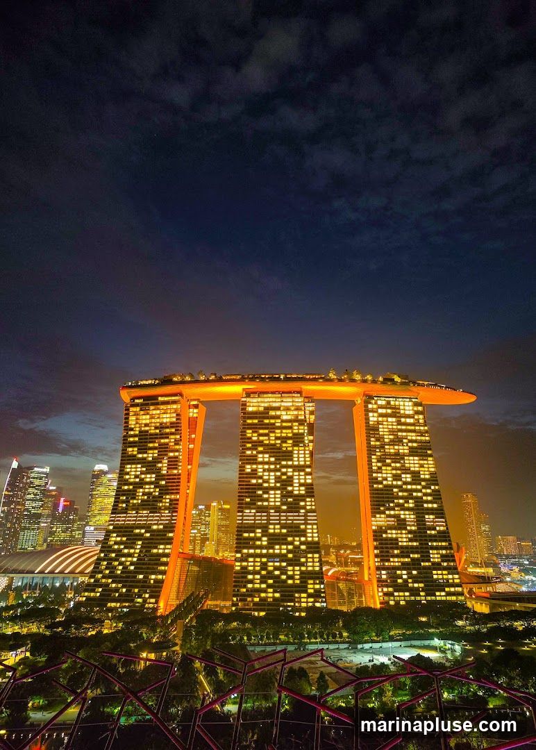 A nighttime view of the Marina Bay Sands hotel in Singapore, with its iconic three towers connected by a sky park. The building is illuminated with orange lights, and the windows of the towers are lit up, creating a grid pattern. The surrounding cityscape is also visible, with other skyscrapers lit up in the distance. The sky is dark blue with some clouds. In the foreground, there are some illuminated structures that appear to be part of a garden or park.