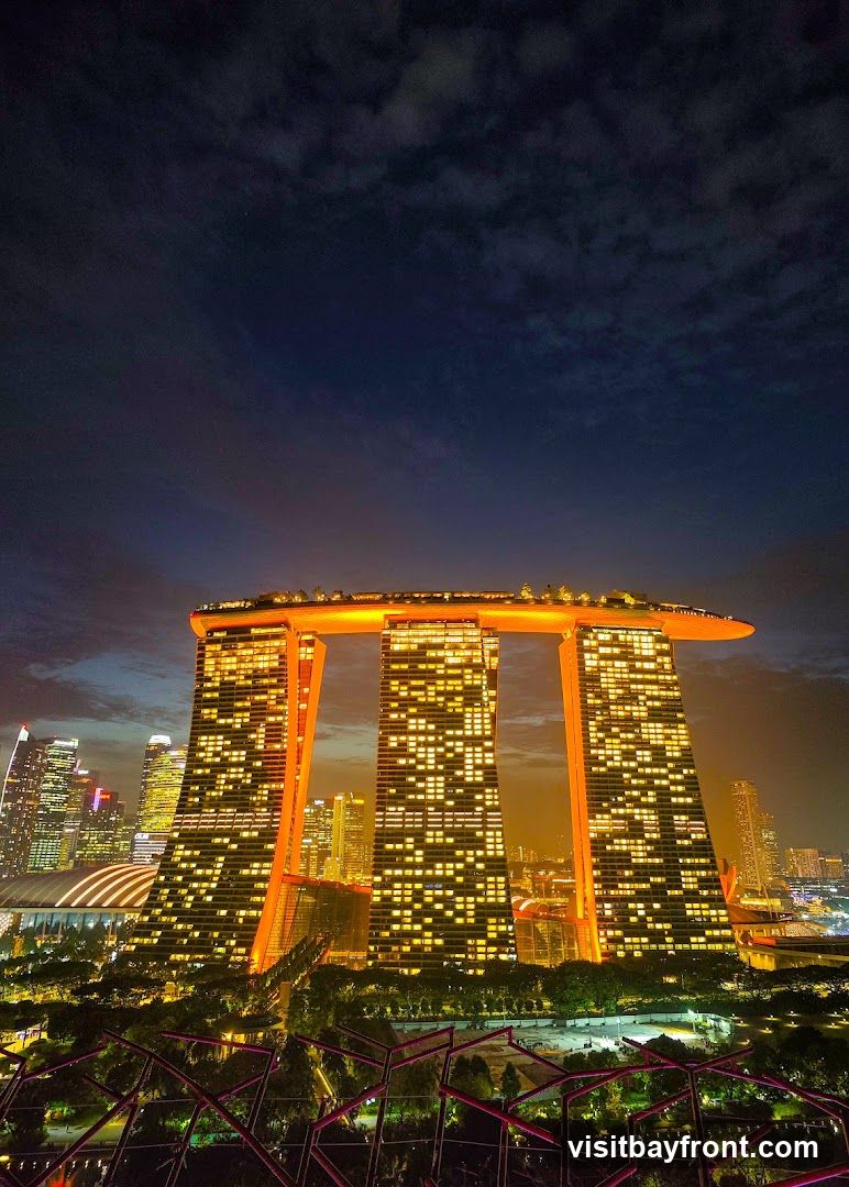 A nighttime view of the Marina Bay Sands hotel in Singapore, with its iconic three towers connected by a sky park. The building is illuminated with orange lights, and the windows of the towers are lit up, creating a grid pattern. The surrounding cityscape is also visible, with other skyscrapers lit up in the distance. The sky is dark blue with some clouds. In the foreground, there are some illuminated structures that appear to be part of a garden or park.