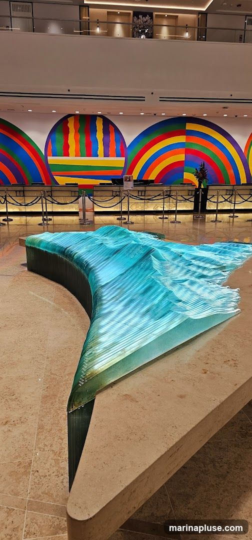 A modern lobby features a striking, wave-like sculpture made of layered blue glass in the foreground. Behind it, a reception desk area is visible with a colorful, abstract mural adorning the wall, depicting semicircles with rainbow and striped patterns. The overall ambiance is contemporary and artistic.