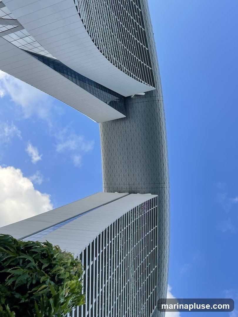 A low-angle shot of a modern architectural complex against a bright blue sky with scattered clouds. The buildings feature sleek, curved designs with a combination of glass and metallic facades. A lush green plant is visible in the foreground.