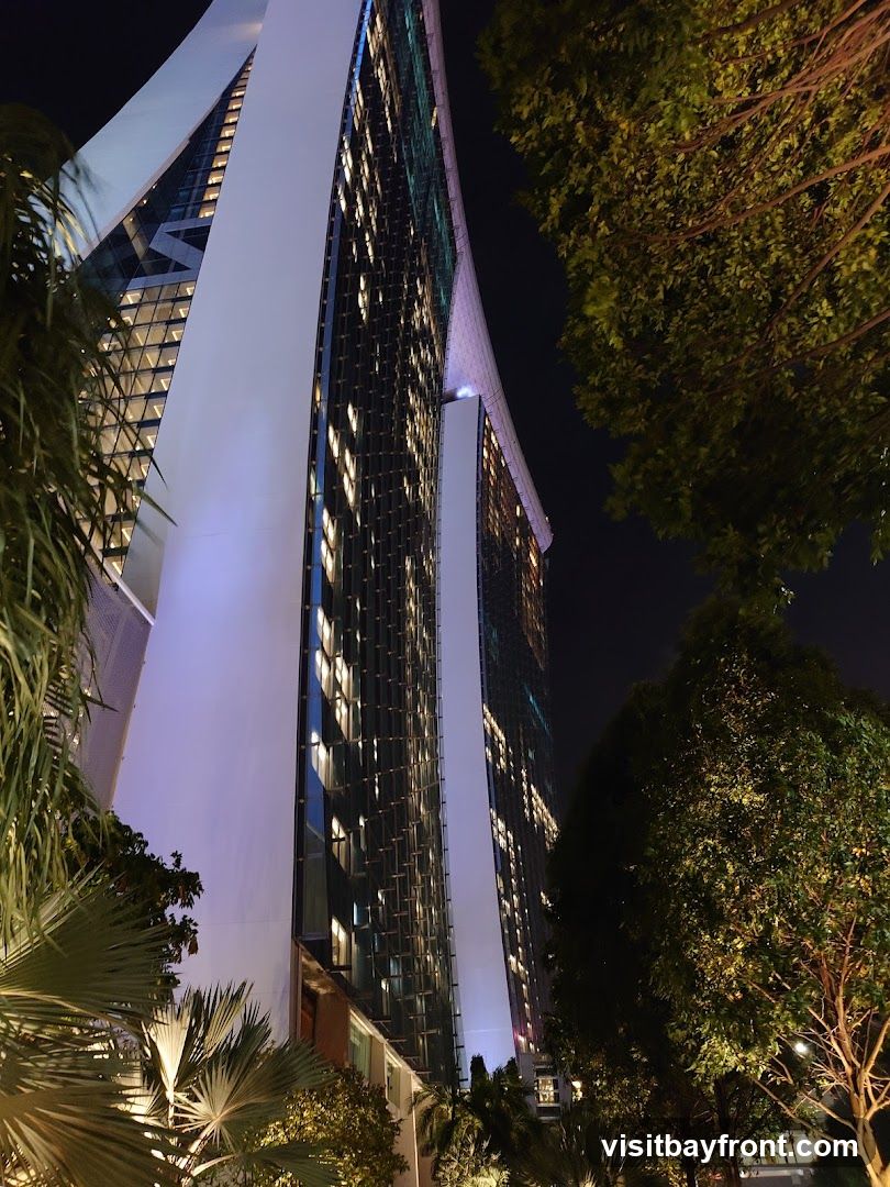 A low-angle shot of a modern skyscraper at night, illuminated by interior lights and accent lighting. Lush green trees and palm fronds frame the building, creating a contrast between nature and architecture. The sky is dark, highlighting the illuminated windows and the sleek design of the building.