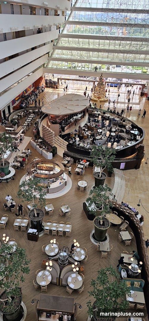 An aerial view of a large, multi-level atrium with a central restaurant and bar area. The space is filled with tables and chairs, some set for dining and others arranged in lounge seating. Potted trees are strategically placed throughout the area, adding a touch of greenery. In the background, a large Christmas tree is visible, suggesting a festive season. People are seen dining, walking, and socializing in the open space. The architecture features a modern design with glass ceilings and multiple balconies overlooking the main floor.