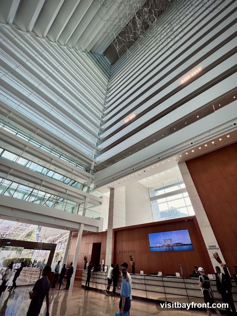 A wide-angle shot of a grand hotel lobby with a high ceiling and multiple floors visible. The architecture features a modern design with clean lines, glass, and wood accents. A large screen displays an image of a cityscape. People are seen interacting at a concierge desk.