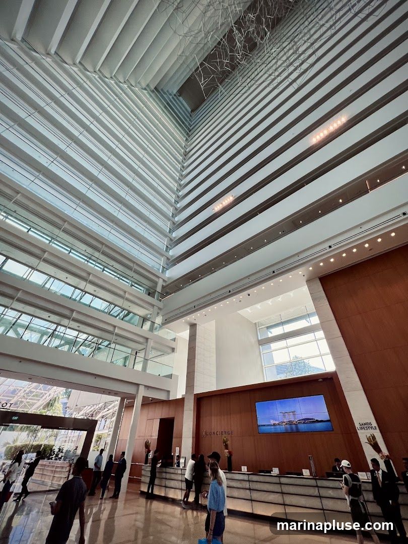 A wide-angle shot of a grand hotel lobby with a high ceiling and multiple floors visible. The architecture features a modern design with clean lines, glass, and wood accents. A large screen displays an image of a cityscape. People are seen interacting at a concierge desk.