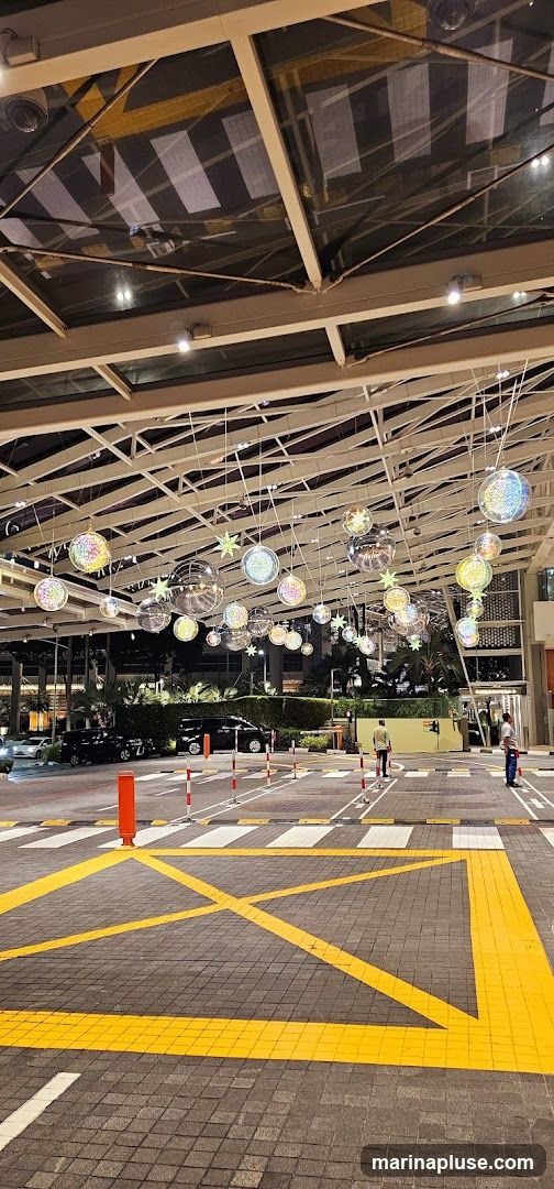 An outdoor walkway at night, featuring a modern roof structure with decorative lights hanging from it. The lights are a mix of disco balls and star shapes, illuminated with colorful lights. The ground is paved with interlocking stones and marked with yellow and white lines, indicating a pedestrian crossing and a no-parking zone. Several cars are parked in the background, and two individuals are visible walking in the distance.