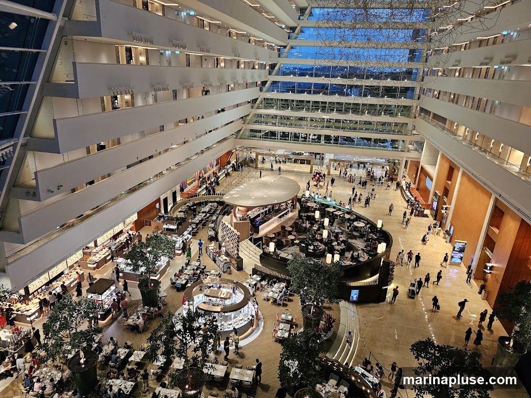 An aerial view of a large, multi-level atrium filled with people dining and walking. Several restaurants and seating areas are visible, with tables set for meals. Lush greenery is interspersed throughout the space. The architecture features tiered levels with balconies overlooking the central area, and a large, abstract sculpture hangs from the ceiling. The overall impression is one of a grand, bustling indoor public space, possibly a hotel lobby or a shopping mall.