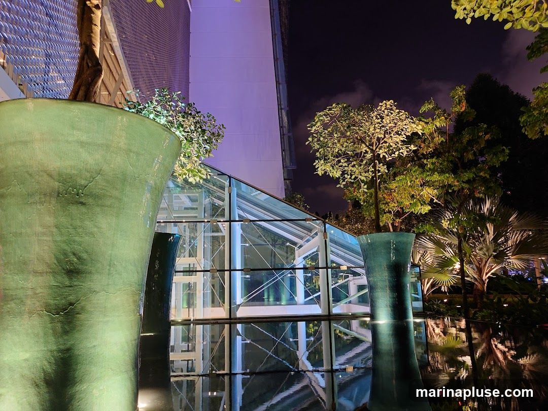 A modern greenhouse structure with glass walls and a metal frame is illuminated at night. Large, green, vase-like planters with trees and foliage are placed in the foreground, with their reflections visible in the water below. The scene is lit with artificial lights, creating a dramatic and serene atmosphere.