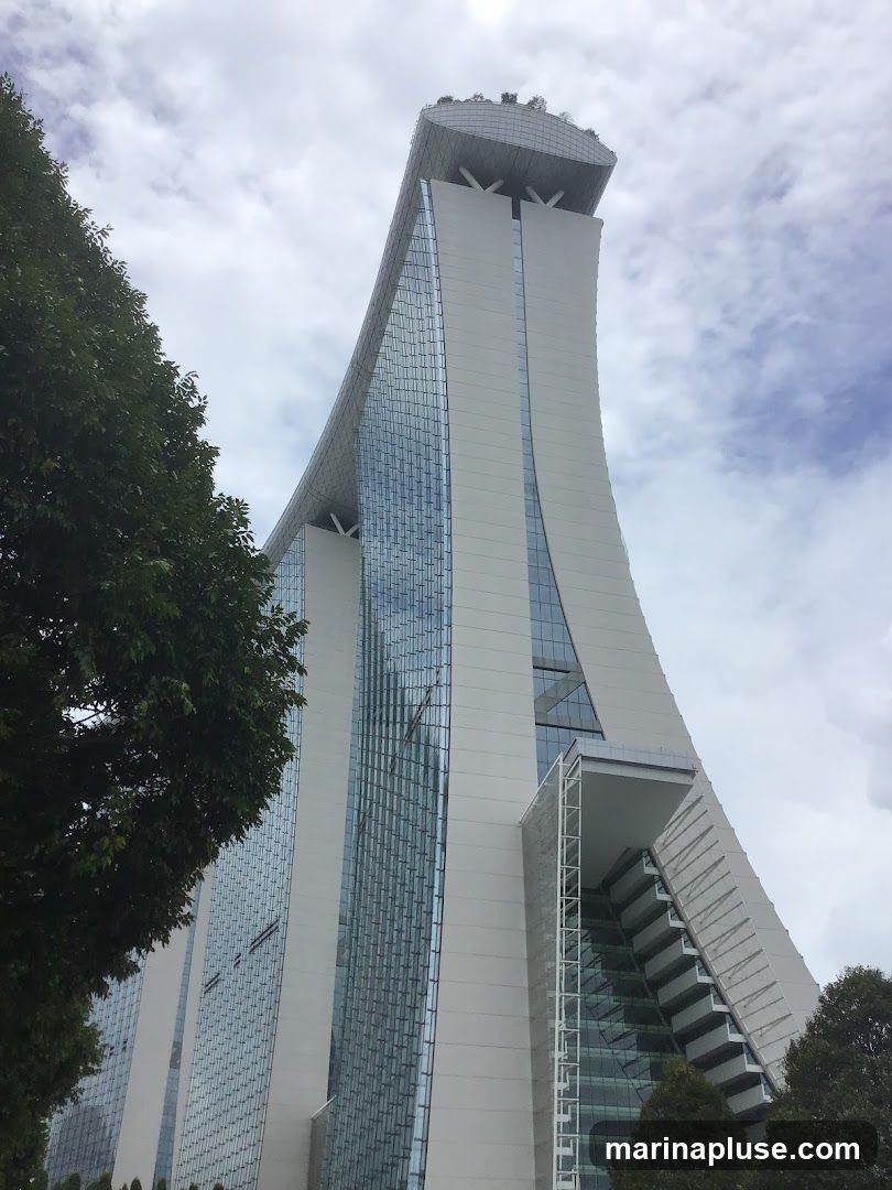 A low-angle shot of the Marina Bay Sands hotel in Singapore, with its distinctive boat-shaped top. The building features a modern design with a white facade and large glass panels. Trees are visible in the foreground and on the rooftop. The sky is cloudy.