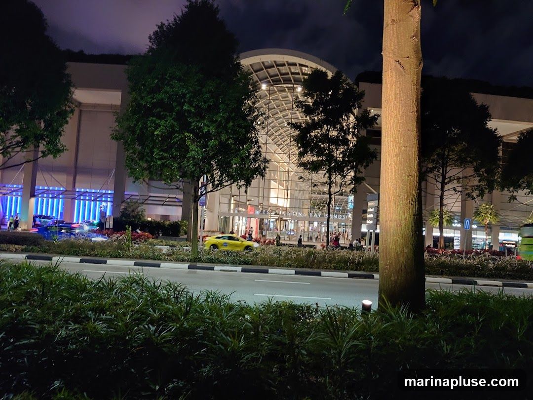 A nighttime view of the Marina Bay Sands complex, with its distinctive architecture illuminated. A yellow taxi is visible on the road in front of the building. Trees and greenery line the foreground and median of the road.