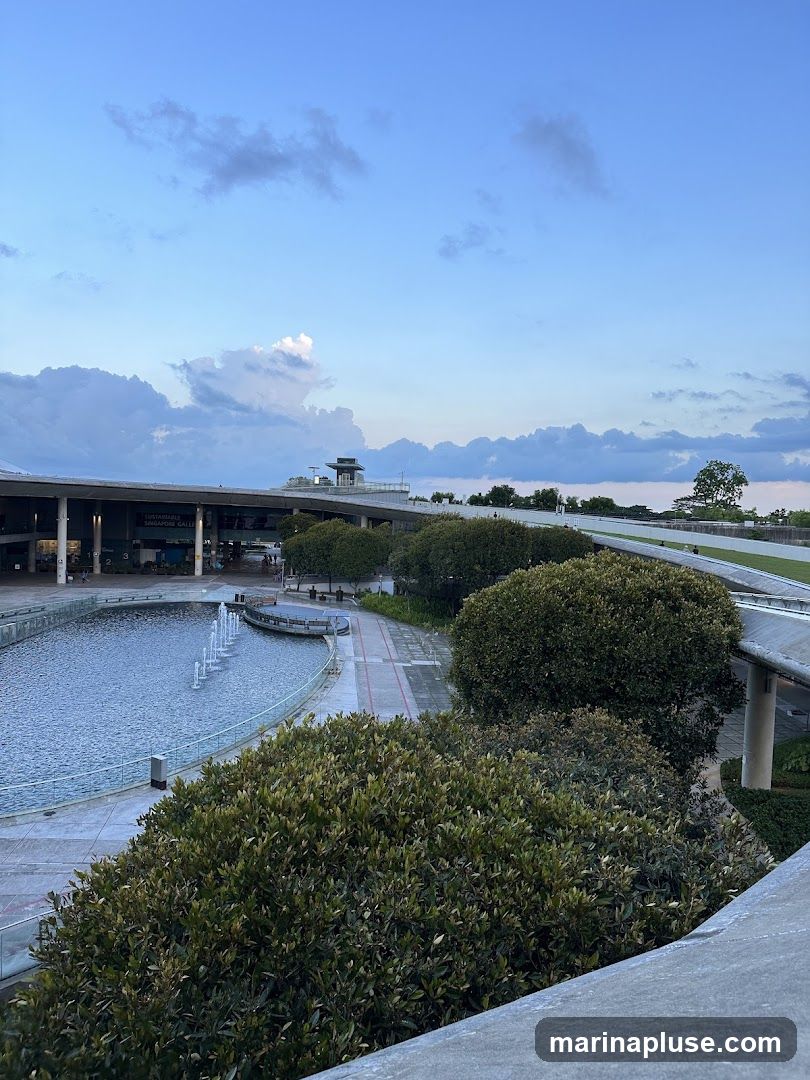 An outdoor plaza with a long fountain in a body of water. Lush green bushes and trees are scattered throughout the scene. In the background, a modern building with a covered walkway is visible under a cloudy blue sky. The building has signage that reads 'SUSTAINABLE SINGAPORE GALLERY'.