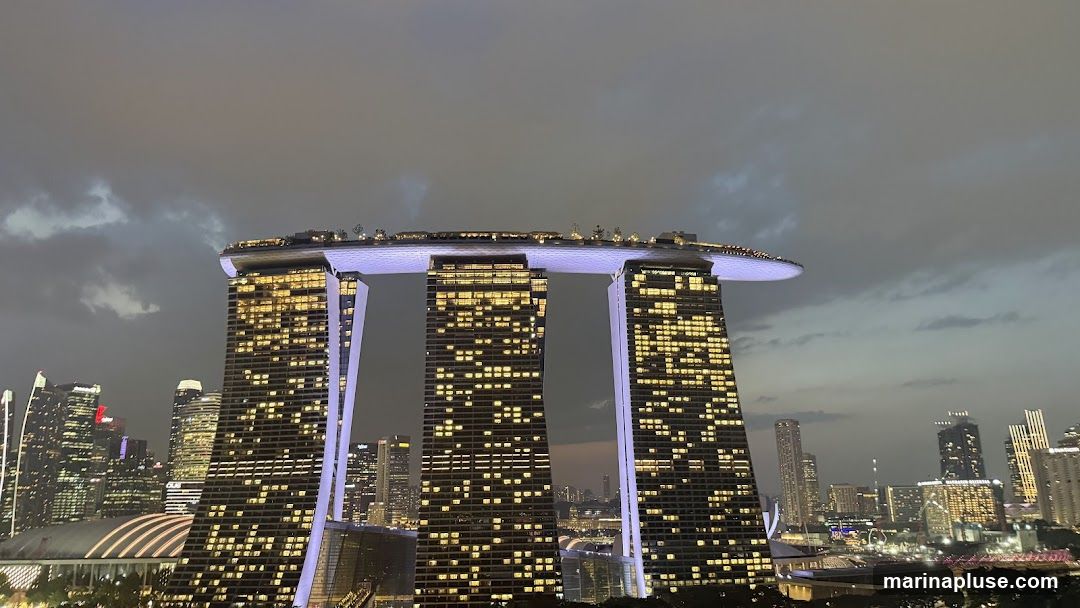 The iconic Marina Bay Sands hotel in Singapore is illuminated against a cloudy dusk sky. The three towers are connected by a large, boat-shaped structure at the top, which is lit with purple lights. Many windows in the towers are lit with warm yellow light, suggesting occupancy. The surrounding cityscape is also visible, with numerous other buildings lit up.