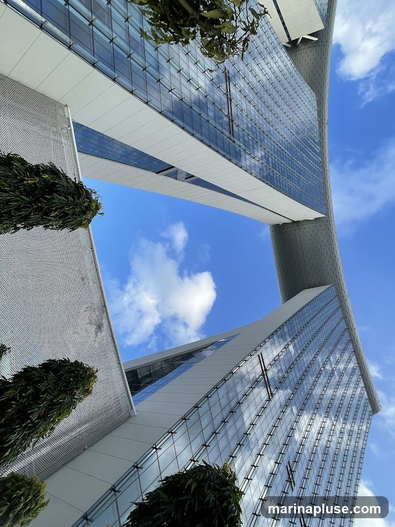 A low-angle shot of a modern building with a glass facade, reflecting the blue sky and clouds. The building features clean lines and geometric shapes, with sections of white paneling. Lush green trees are visible in the foreground, adding a touch of nature to the urban landscape.