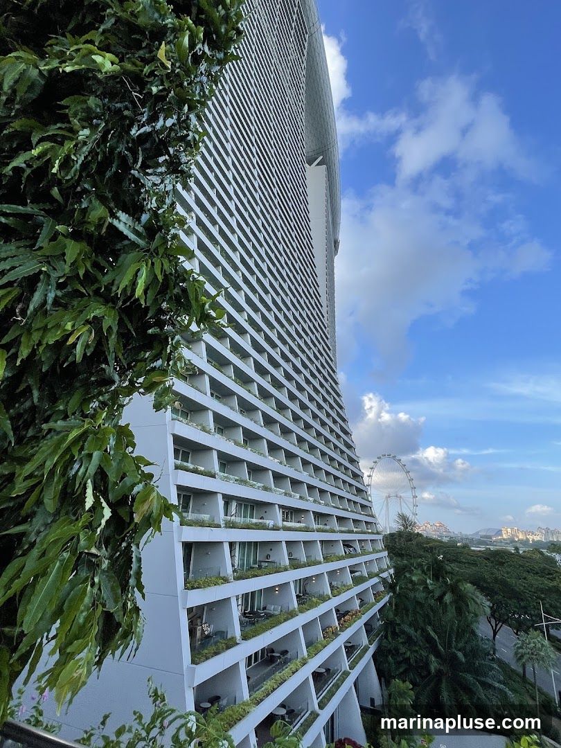A tall, modern skyscraper with a distinctive curved facade is shown from a low angle, with lush green foliage in the foreground. The building features numerous balconies, many with outdoor seating and plants, suggesting residential or hotel use. In the background, a Ferris wheel and a cityscape are visible under a partly cloudy blue sky. The architecture emphasizes clean lines and a sense of scale.