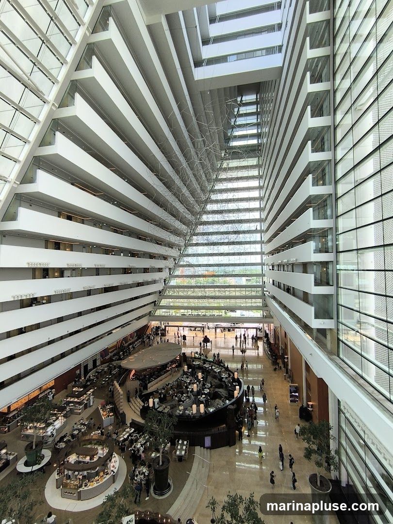 An interior view of a large, modern hotel atrium with multiple levels of balconies overlooking a central dining area. The architecture features a striking geometric design with white horizontal lines and glass facades. People are seen walking and dining in the open space below.
