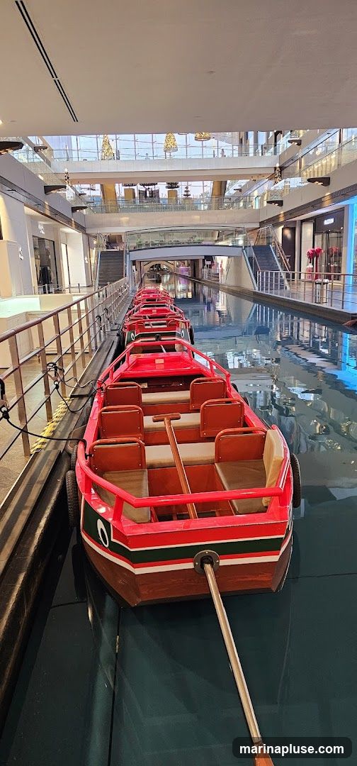 A row of red gondola boats are docked in a canal that runs through a modern shopping mall. The boats have wooden seats and are equipped with oars. The mall features multiple levels with shops and escalators visible in the background. Decorative Christmas trees are also visible on an upper level.