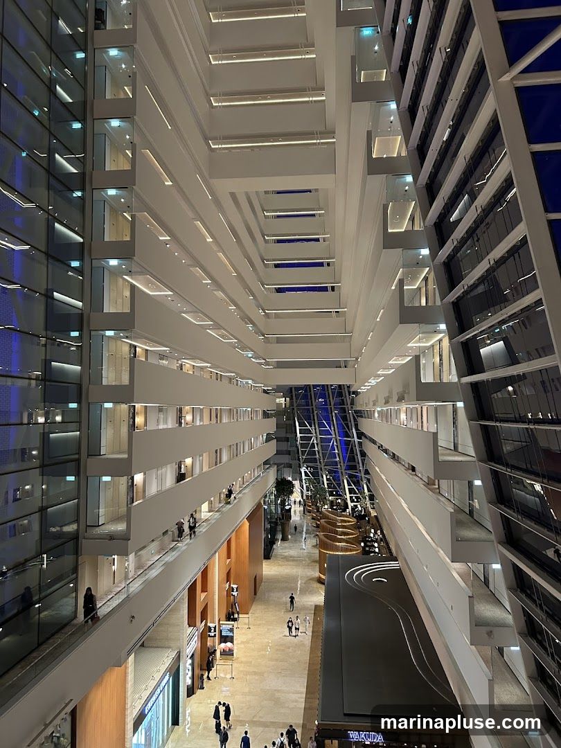 The image shows a view looking down into a vast atrium of a modern building. Multiple levels of walkways and rooms are visible, with glass facades on one side and white balconies on the other. People are seen walking on the lower levels and on the walkways. The atrium has a large, angled glass ceiling with a blue hue. There are some plants and seating areas visible on the ground floor.