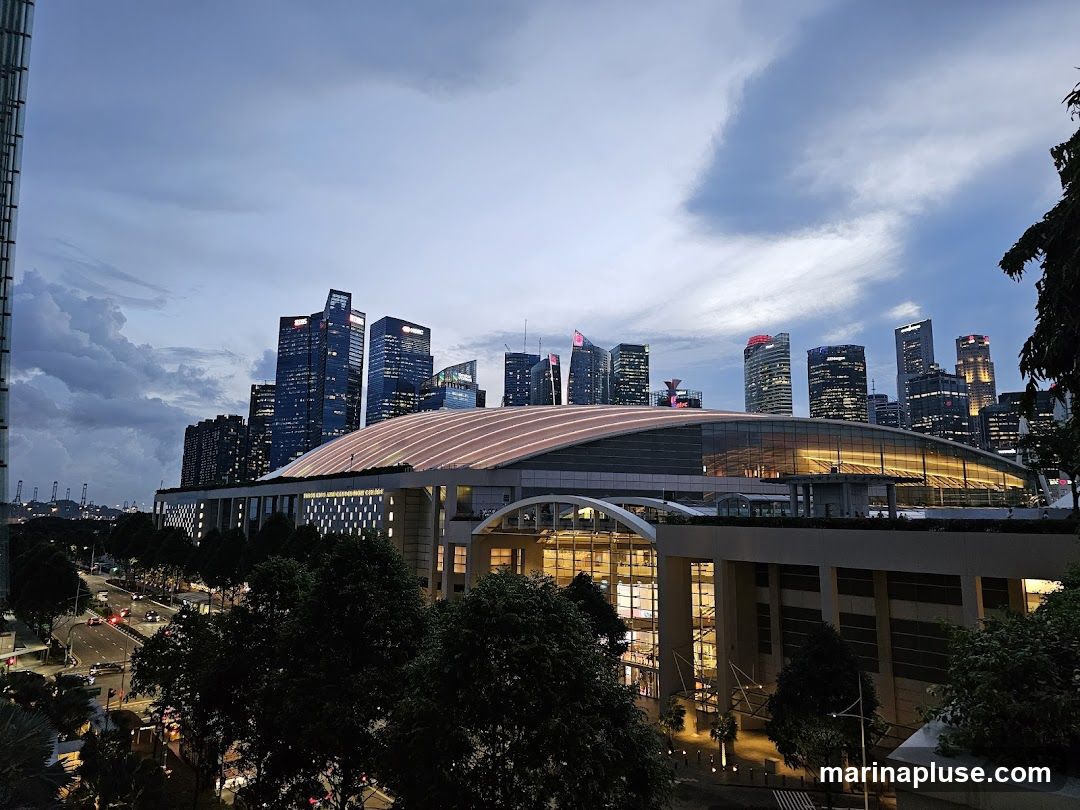 A panoramic view of a modern cityscape at dusk, featuring a large domed building illuminated with warm lights, surrounded by numerous skyscrapers. The sky is a mix of deep blue and dramatic clouds, with the city lights beginning to twinkle. Trees and a road with traffic are visible in the foreground, adding depth to the scene.