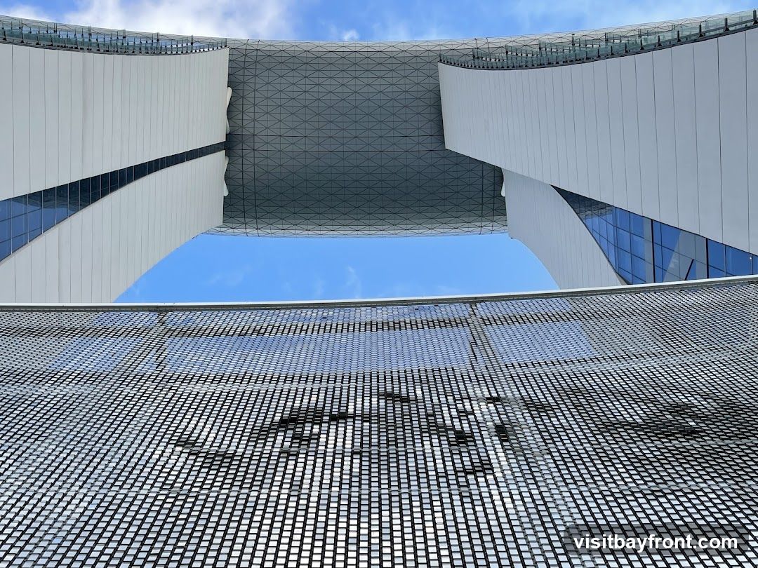 A low-angle shot of a modern architectural structure against a bright blue sky with scattered clouds. The building features curved white panels and glass windows, with a geometric patterned roof or ceiling visible. In the foreground, a metallic mesh or grate creates a textured foreground element.