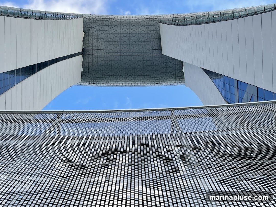 A low-angle shot of a modern architectural structure against a bright blue sky with scattered clouds. The building features curved white panels and glass windows, with a geometric patterned roof or ceiling visible. In the foreground, a metallic mesh or grate creates a textured foreground element.