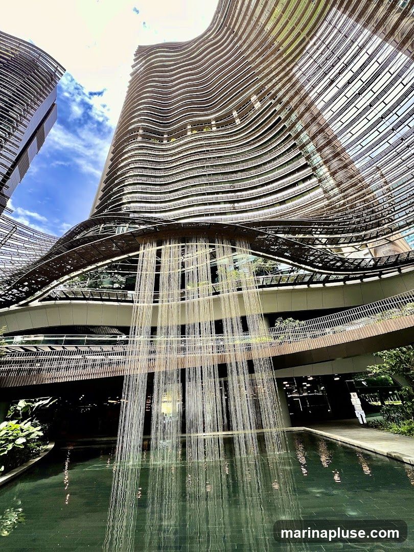 A low-angle shot of a modern, curved skyscraper with a unique facade. A water feature cascades down from an elevated walkway into a pool below, reflecting the surrounding architecture and greenery. The sky is visible with scattered clouds.