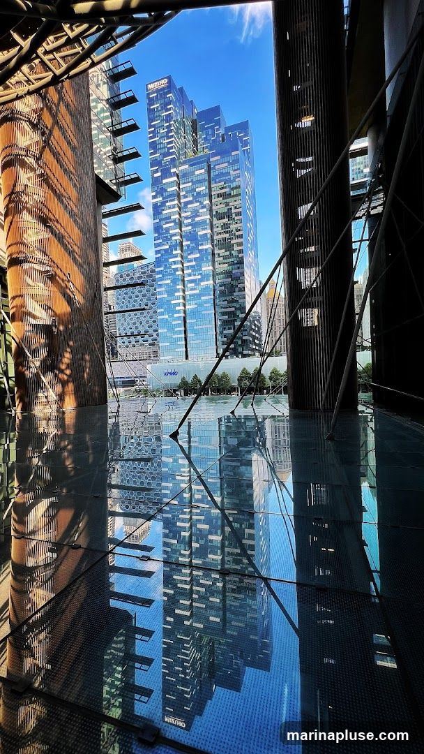 A view through a modern architectural structure, showcasing the reflections of tall, glass-facade buildings on a reflective surface. The sky is bright blue with scattered clouds. The buildings feature intricate patterns of windows and some have visible signage, including 'MIZUHO' and 'KPMG'. The foreground is dominated by the geometric patterns of the reflective surface and the supporting structures of the viewpoint.