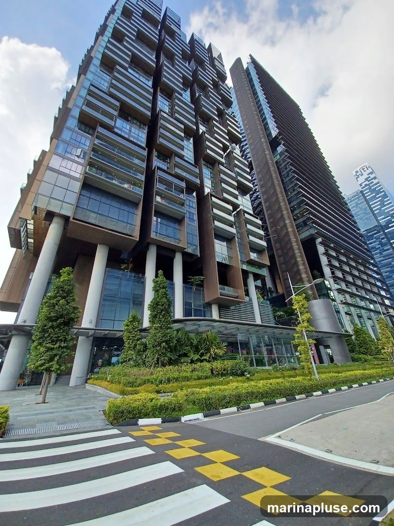 A low-angle shot captures the striking architecture of a modern skyscraper with a unique, tiered design featuring numerous balconies and glass facades. The building is surrounded by meticulously landscaped greenery, including manicured bushes and tall trees, creating a contrast between the urban structure and nature. In the foreground, a pedestrian crossing with yellow and white stripes leads towards the building. The sky above is partly cloudy.