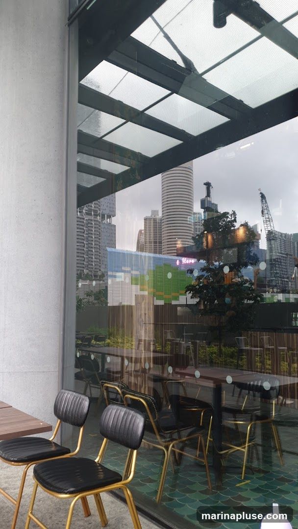 A view of an outdoor seating area with tables and chairs, reflected in a large window. Beyond the seating, a cityscape with modern buildings and construction cranes is visible. A neon sign that reads "t Have" is also reflected in the glass.