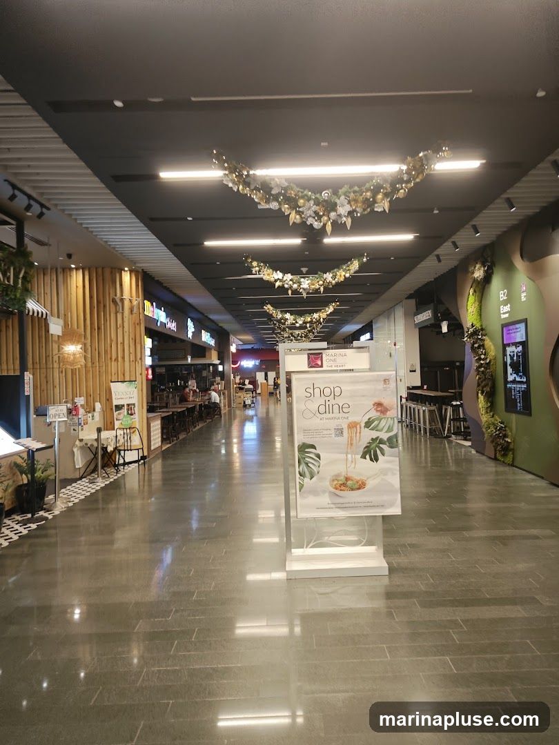 A wide, polished stone corridor inside a modern shopping mall. Festive garlands with lights and ornaments are suspended from the dark ceiling. On the left, a section of shops with bamboo-style paneling is visible, including a sign that says "OPEN". Further down, a promotional poster for "Shop & Dine at Marina One" is displayed on a stand. The corridor leads to a food court area with tables and chairs.