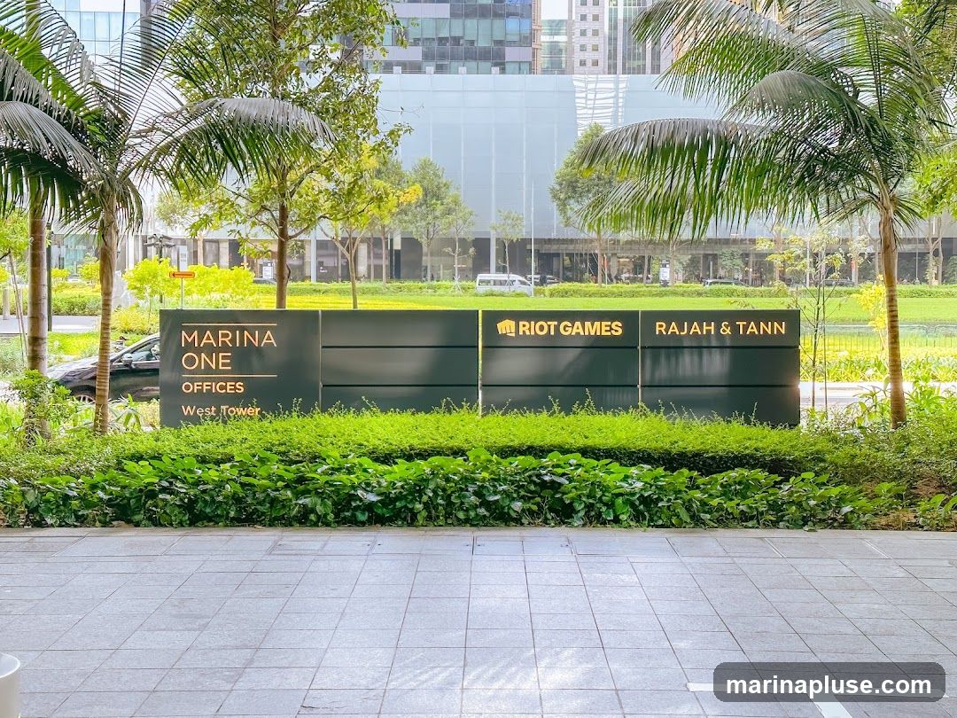 A dark green sign with illuminated text indicates "MARINA ONE OFFICES West Tower" on the left, and "RIOT GAMES" and "RAJAH & TANN" on the right. The sign is surrounded by lush green landscaping and palm trees, with modern office buildings visible in the background.