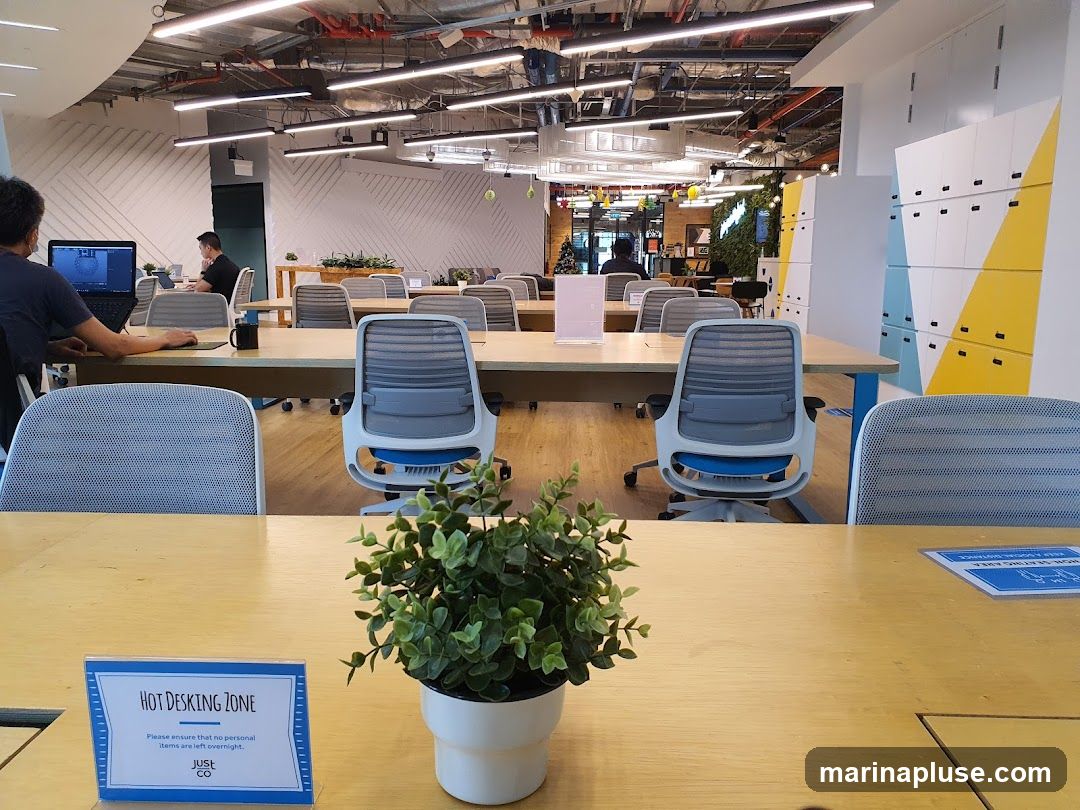 An interior view of a modern co-working space. The image shows rows of desks with office chairs, some occupied by individuals working on laptops. In the foreground, a sign indicates a "HOT DESKING ZONE" with instructions to remove personal items overnight. A small potted plant sits on a desk. In the background, there are lockers and a green wall with plants. The ceiling features exposed industrial elements and lighting fixtures.