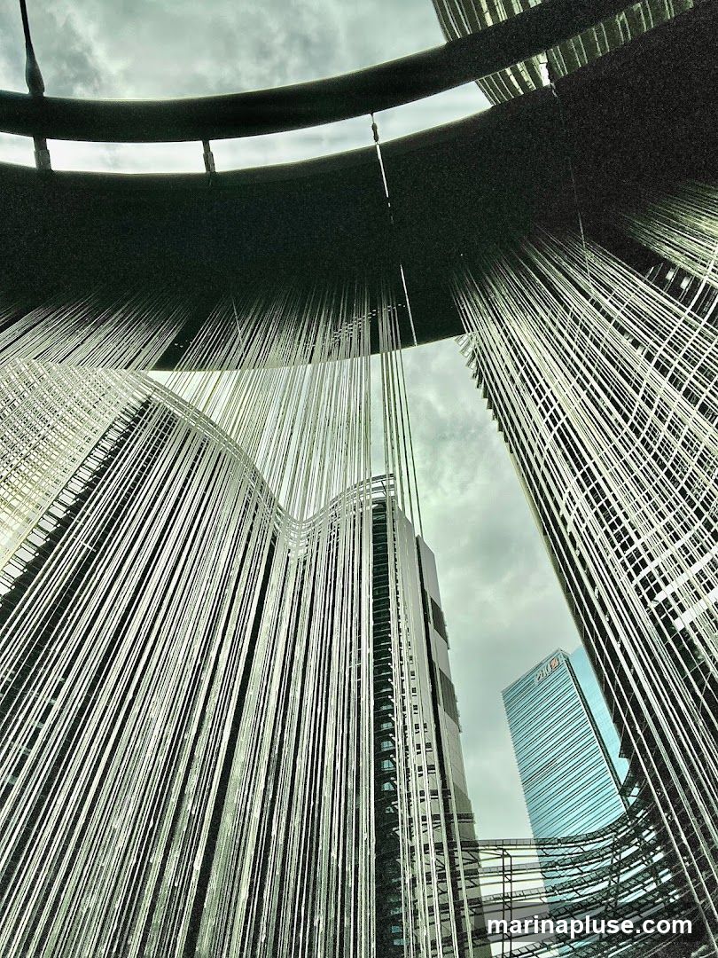 A low-angle shot looking up at modern skyscrapers partially obscured by a contemporary art installation. The installation consists of numerous thin, reflective strands hanging from a dark, curved structure, creating a curtain-like effect. The strands reflect the sky and the surrounding buildings. One building in the background has a blue facade with the logo 'PMI' visible. The sky is overcast with dramatic clouds.