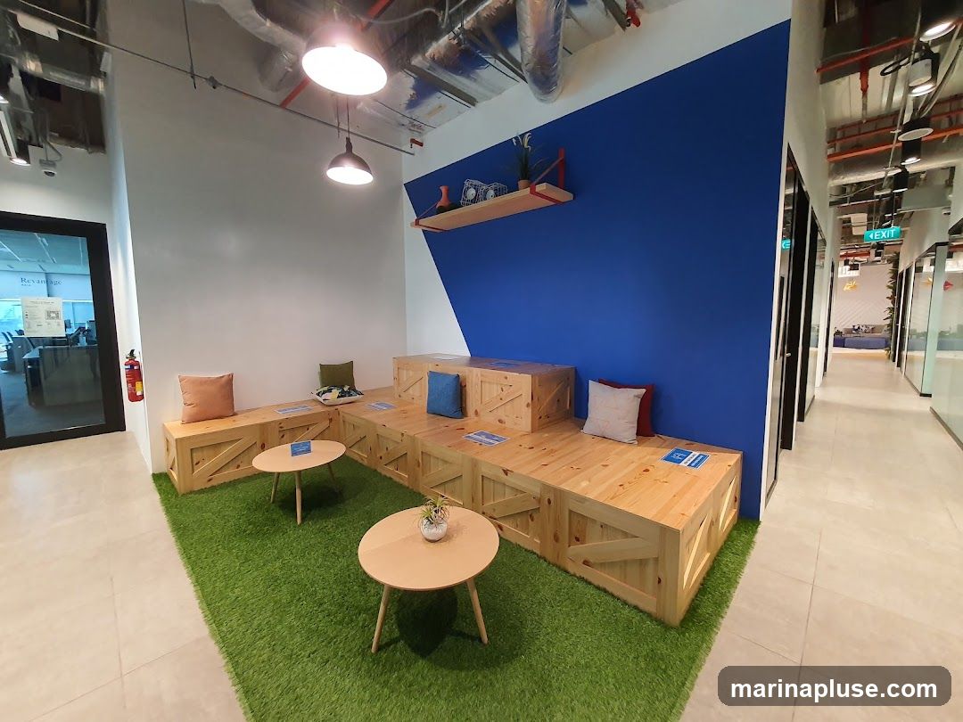 A contemporary office break area features a built-in wooden bench seating arrangement with colorful cushions, set against a vibrant blue accent wall. Two light wood coffee tables are placed on a patch of artificial grass, creating a relaxed and inviting atmosphere. The space is well-lit with overhead industrial-style lighting. A hallway with glass-walled offices is visible to the right, and a door with a sign indicating "Revantage" is on the left.