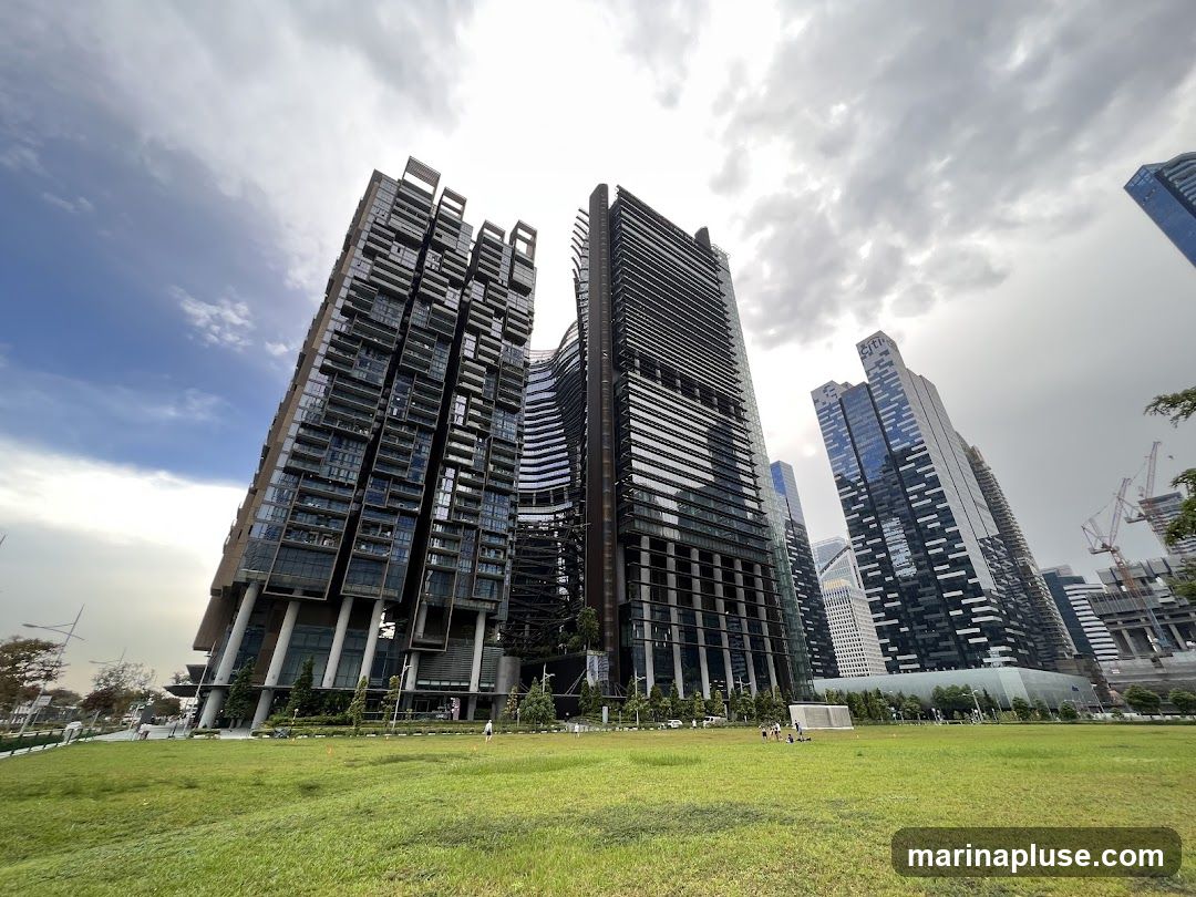 A wide-angle shot captures a cluster of modern skyscrapers with unique architectural designs, set against a cloudy sky. In the foreground, a lush green lawn stretches out, with a few people visible in the distance, some appearing to be playing on the field. The buildings are tall and sleek, featuring glass facades and intricate structural elements. The overall scene depicts a blend of urban development and open green space.