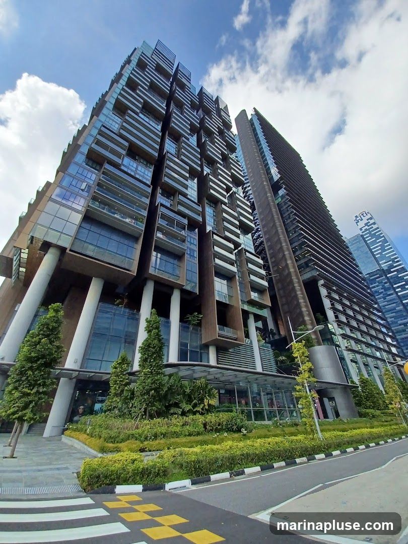 A low-angle shot of a modern, multi-story building with a unique architectural design featuring balconies and glass facades. The building is surrounded by lush greenery, including trees and manicured bushes. In the background, other tall skyscrapers are visible under a partly cloudy sky. A pedestrian crossing with yellow and white markings is in the foreground.