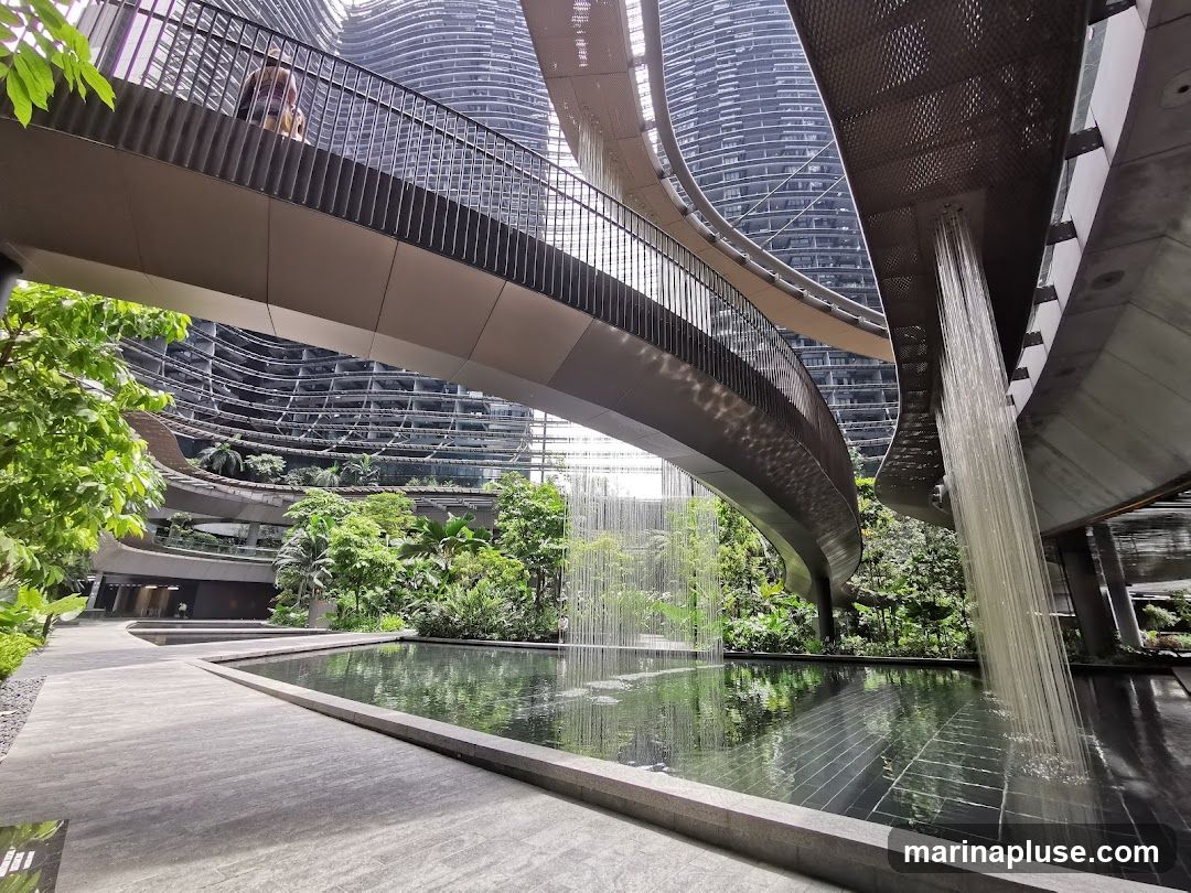 An interior view of a modern atrium featuring curved walkways, lush greenery, and water features. A person is visible on an elevated walkway. Water cascades down from above into a reflective pool in the foreground. The architecture is sleek and contemporary, with curved building facades in the background.