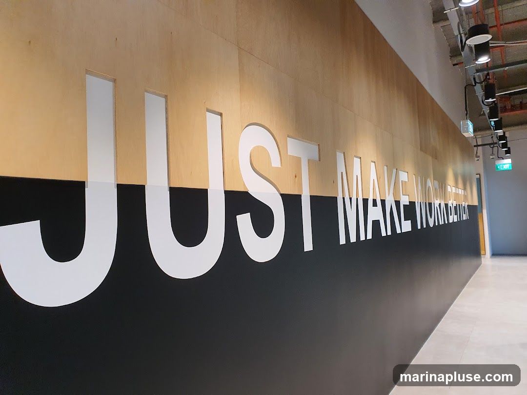 A large wall decal with white lettering on a wood-paneled wall reads "JUST MAKE WORK BETTER". The lettering is cut out and appears to be mounted on the wall. Below the wood paneling is a black section of the wall. The image shows a hallway with lighting fixtures on the ceiling and an exit sign visible in the distance.